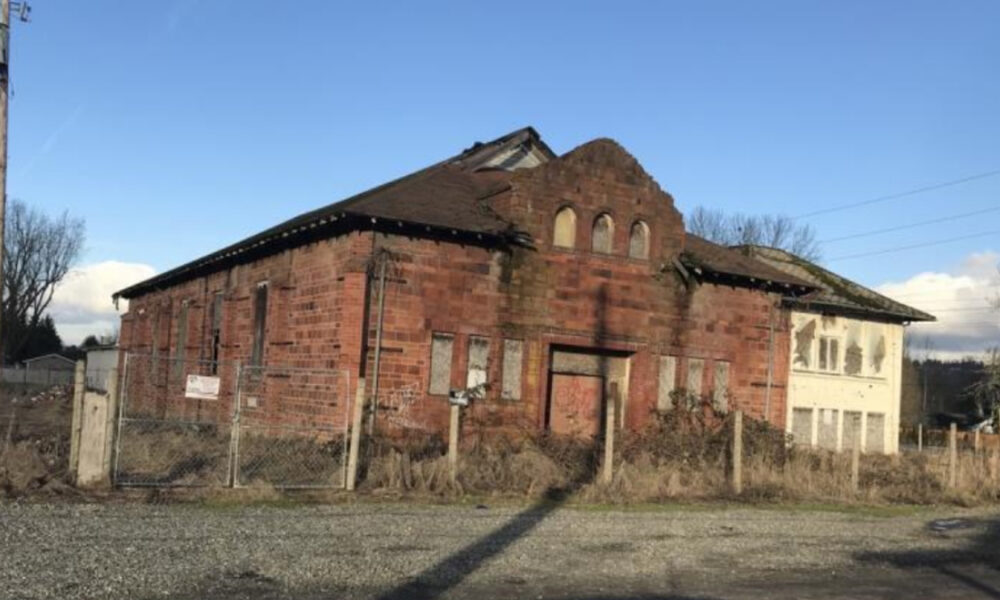 Abandoned red brick building with arched windows and overgrown yard, fenced in a rural lot under a clear blue sky.