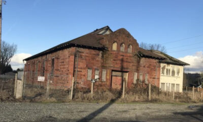 Abandoned red brick building with arched windows and overgrown yard, fenced in a rural lot under a clear blue sky.