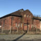 Abandoned red brick building with arched windows and overgrown yard, fenced in a rural lot under a clear blue sky.