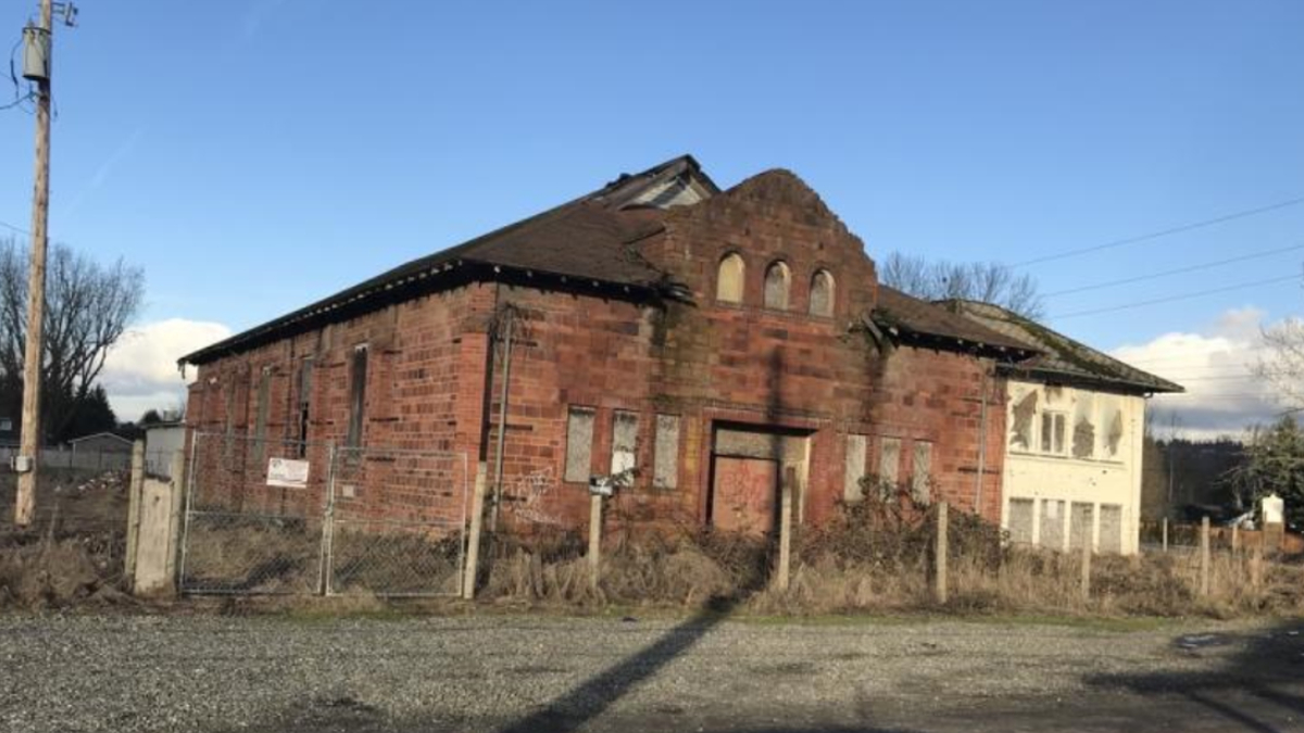 Abandoned red brick building with arched windows and overgrown yard, fenced in a rural lot under a clear blue sky.