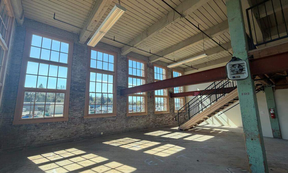 Sunlit industrial loft with brick walls, tall sash windows, and a metal staircase to a mezzanine.