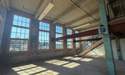 Sunlit industrial loft with brick walls, tall sash windows, and a metal staircase to a mezzanine.