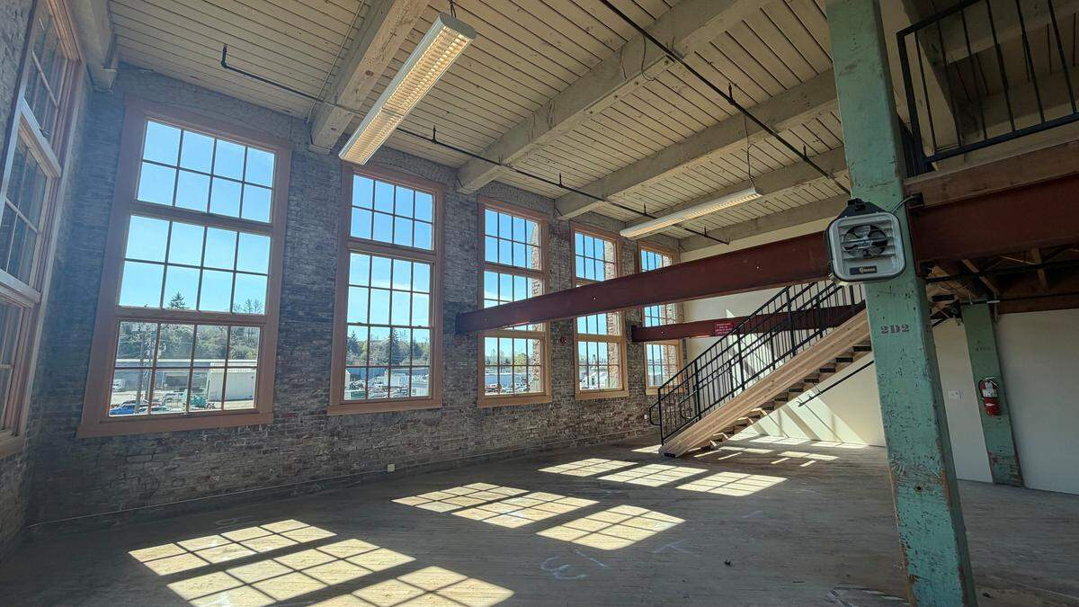 Sunlit industrial loft with brick walls, tall sash windows, and a metal staircase to a mezzanine.
