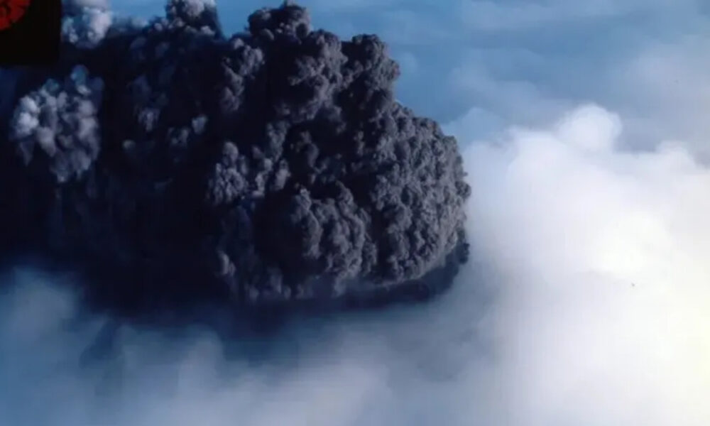 Dark, dense volcanic ash plume rising from an eruption, with surrounding blue sky and light mist at the base