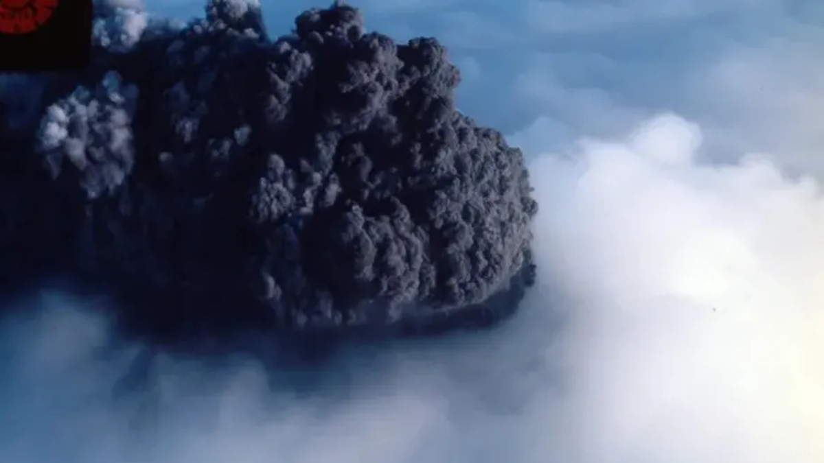 Dark, dense volcanic ash plume rising from an eruption, with surrounding blue sky and light mist at the base