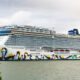 Large cruise ship docked at a harbor with a bright multicolored wave mural along the hull.