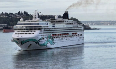 Large white cruise ship with teal wave artwork on bow sailing in a calm harbor, shoreline and houses visible in the background under a cloudy sky.