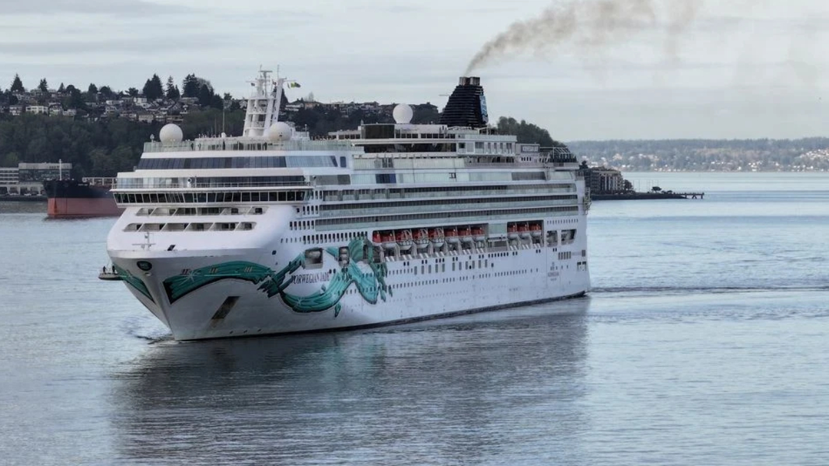 Large white cruise ship with teal wave artwork on bow sailing in a calm harbor, shoreline and houses visible in the background under a cloudy sky.