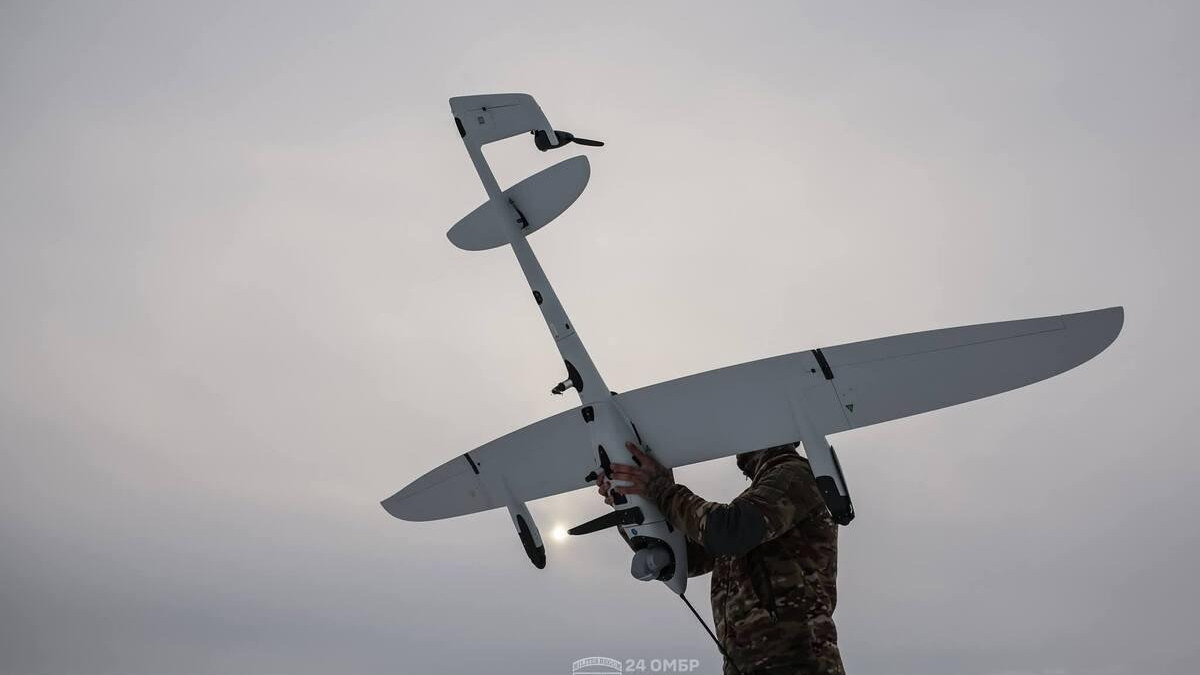 Person in camouflage handles a white unmanned aerial vehicle with a long wing against a cloudy sky.
