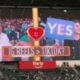 Stadium jumbotron shows two fans holding blue YES signs, a central heart emblem, and the caption 'IG REELS > TIKTOK?'