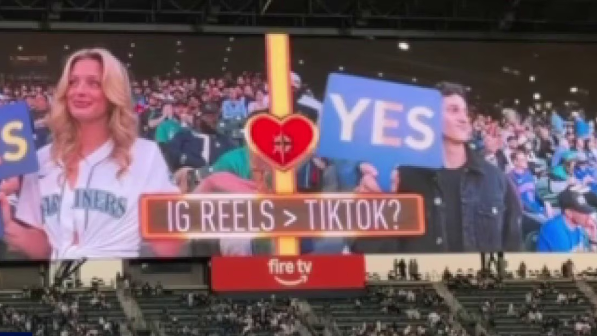 Stadium jumbotron shows two fans holding blue YES signs, a central heart emblem, and the caption 'IG REELS > TIKTOK?'
