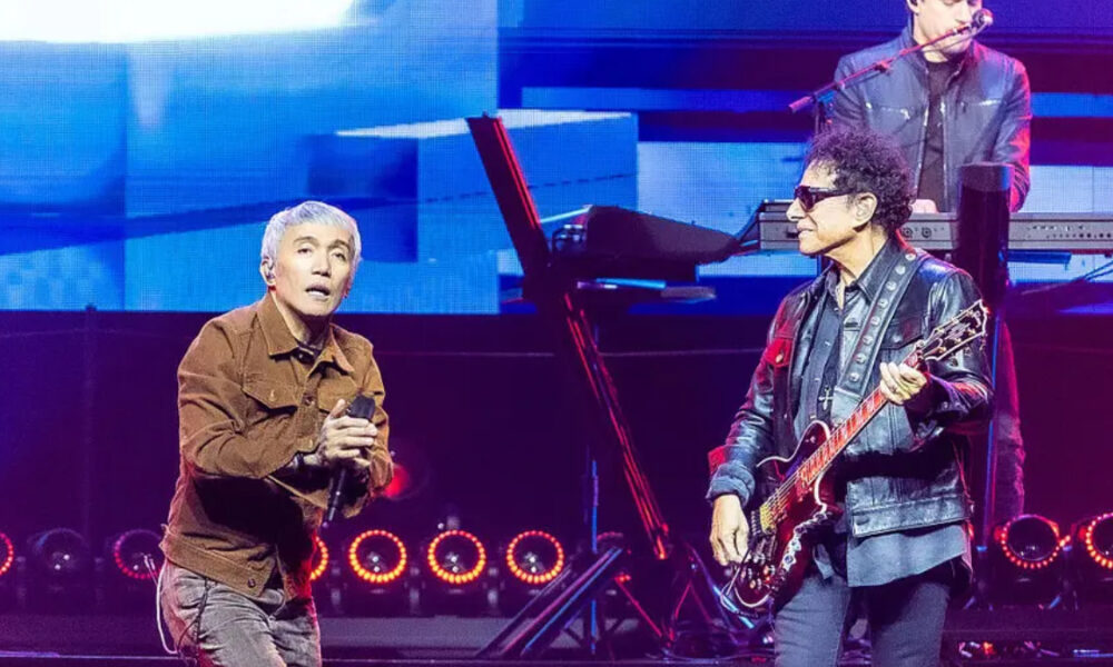 Lead singer with gray hair in a brown jacket holds a microphone while a guitarist in a leather jacket and sunglasses plays beside him on a bright concert stage with a keyboardist in the background.
