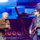Lead singer with gray hair in a brown jacket holds a microphone while a guitarist in a leather jacket and sunglasses plays beside him on a bright concert stage with a keyboardist in the background.