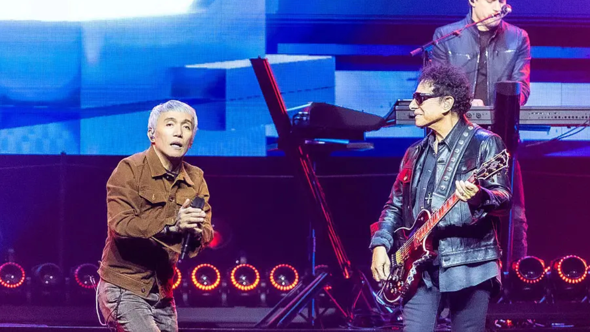 Lead singer with gray hair in a brown jacket holds a microphone while a guitarist in a leather jacket and sunglasses plays beside him on a bright concert stage with a keyboardist in the background.
