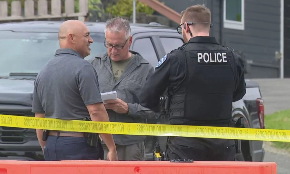 Two men speak with a police officer at a taped-off outdoor scene, one man holding a notebook; a dark pickup and yellow caution tape are in view in front of them.