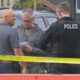 Two men speak with a police officer at a taped-off outdoor scene, one man holding a notebook; a dark pickup and yellow caution tape are in view in front of them.