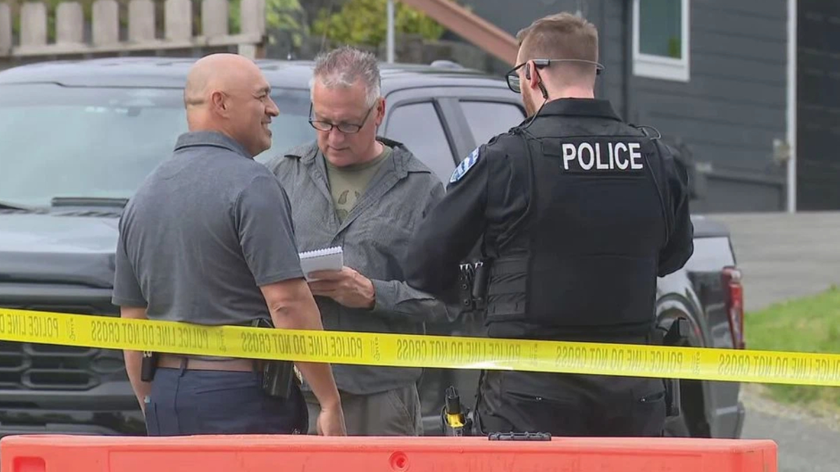 Two men speak with a police officer at a taped-off outdoor scene, one man holding a notebook; a dark pickup and yellow caution tape are in view in front of them.