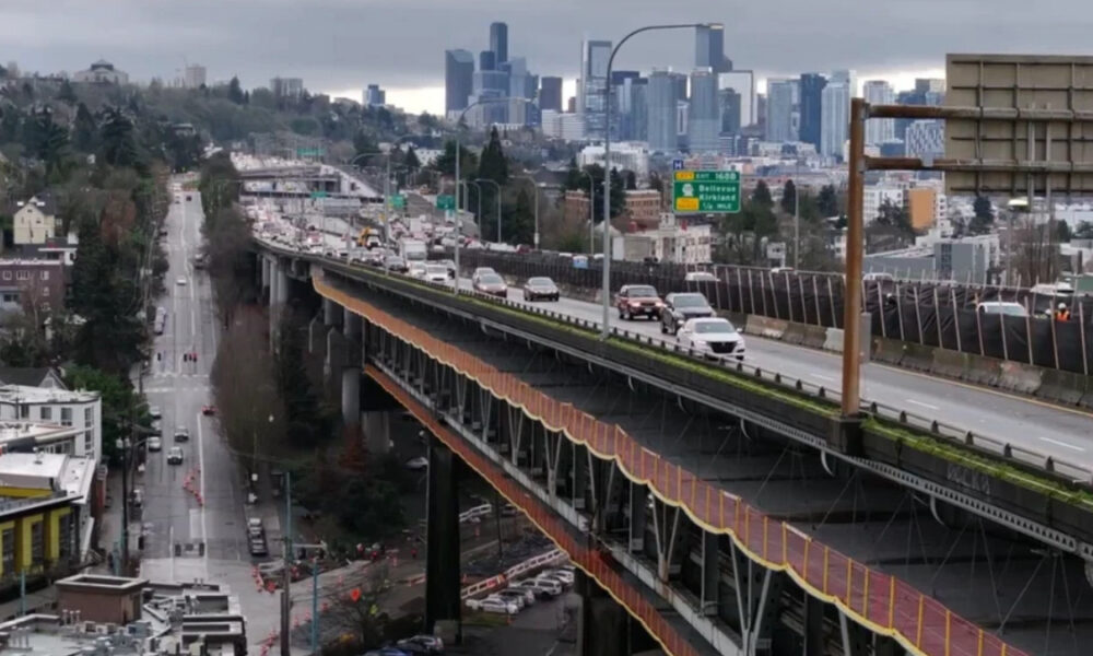 Elevated freeway with cars crossing above a city street, and a distant downtown skyline under a cloudy sky.
