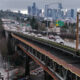 Elevated freeway with cars crossing above a city street, and a distant downtown skyline under a cloudy sky.