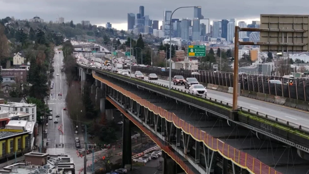 Elevated freeway with cars crossing above a city street, and a distant downtown skyline under a cloudy sky.