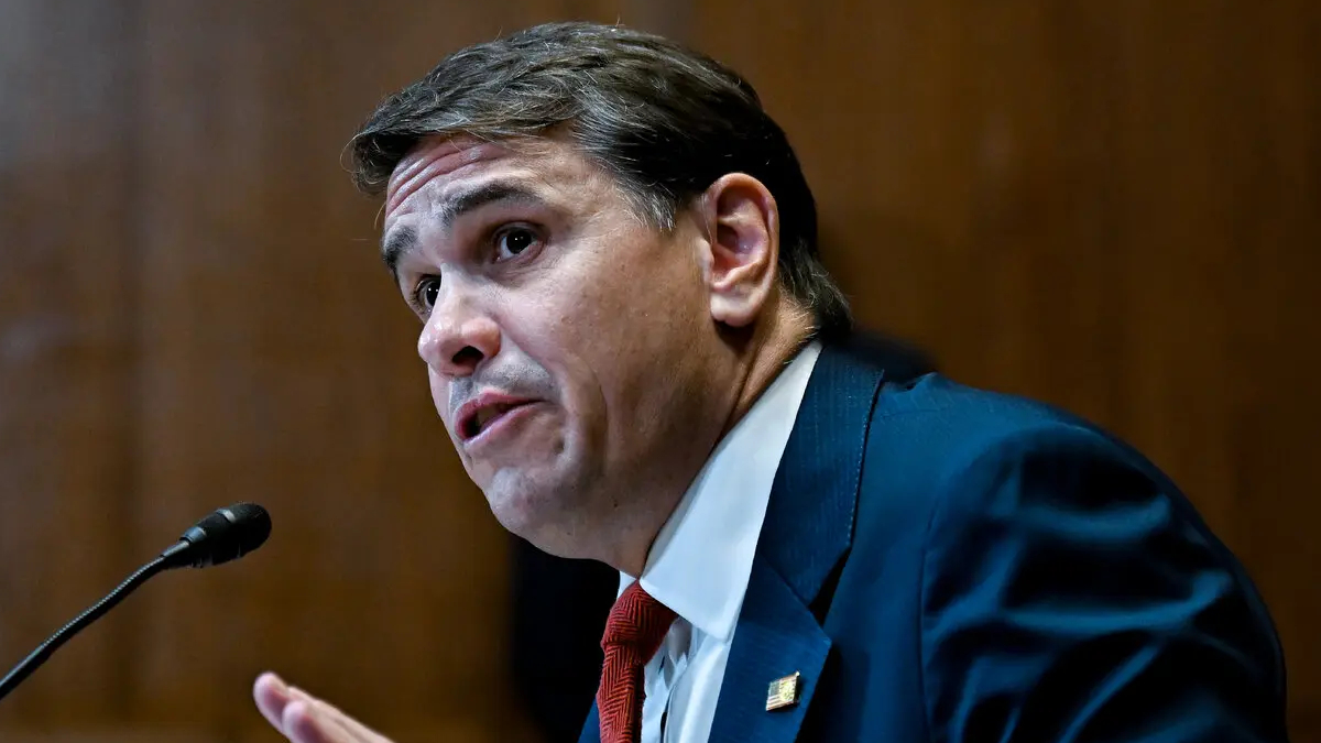 Man in a dark blue suit and red tie speaks into a microphone, mid-speech, against a wooden background.