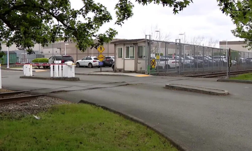 Railroad crossing barriers in front of a gated industrial parking lot with a security booth and fenced perimeter.