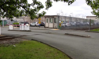 Railroad crossing barriers in front of a gated industrial parking lot with a security booth and fenced perimeter.