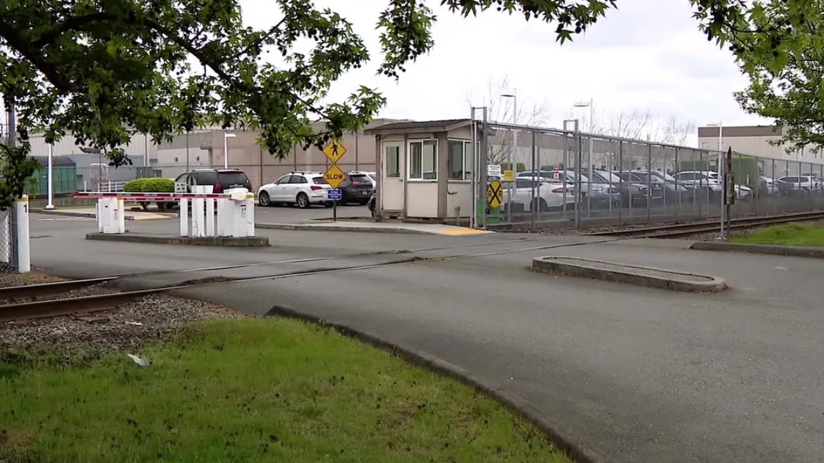 Railroad crossing barriers in front of a gated industrial parking lot with a security booth and fenced perimeter.