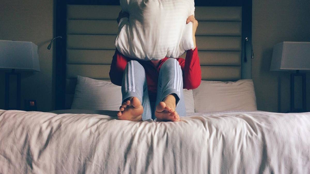 Couple cuddling on a bed, with one person sitting and legs extended toward the camera while the other hugs from behind over a white blanket.