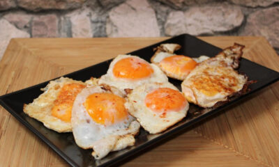 Five sunny-side-up eggs arranged on a black rectangular platter on a wooden table. The eggs have bright orange yolks and slightly browned edges.