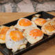 Five sunny-side-up eggs arranged on a black rectangular platter on a wooden table. The eggs have bright orange yolks and slightly browned edges.