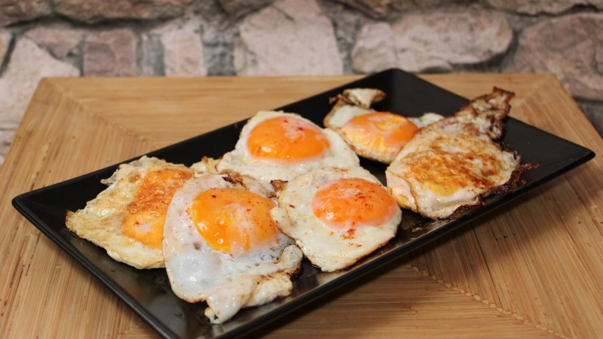 Five sunny-side-up eggs arranged on a black rectangular platter on a wooden table. The eggs have bright orange yolks and slightly browned edges.