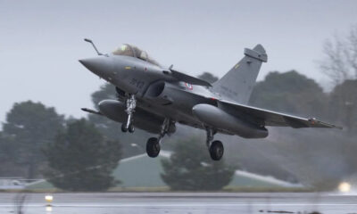 Two-seat military fighter jet lifting off the wet runway in overcast weather, gear still extended.