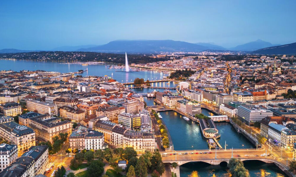 Aerial cityscape of a lakeside city with bridges, a tall water jet, and mountains in the distance at dusk.
