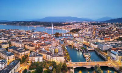 Aerial cityscape of a lakeside city with bridges, a tall water jet, and mountains in the distance at dusk.