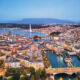 Aerial cityscape of a lakeside city with bridges, a tall water jet, and mountains in the distance at dusk.