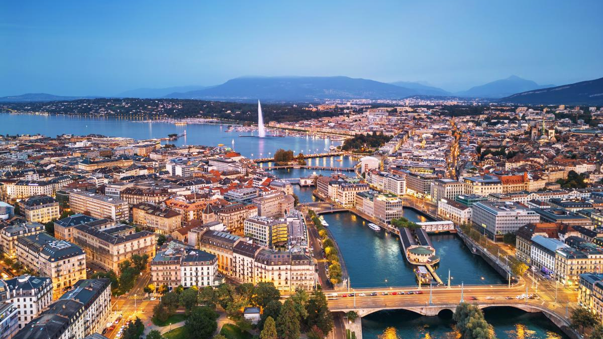 Aerial cityscape of a lakeside city with bridges, a tall water jet, and mountains in the distance at dusk.