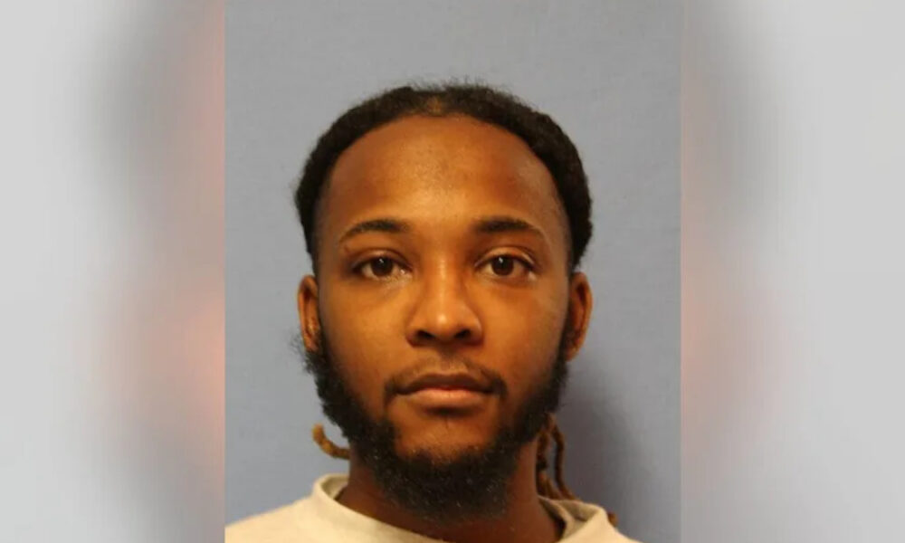 Mugshot-style portrait of a man with short dreadlocks and beard, neutral expression, gray-blue background.