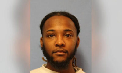 Mugshot-style portrait of a man with short dreadlocks and beard, neutral expression, gray-blue background.