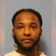 Mugshot-style portrait of a man with short dreadlocks and beard, neutral expression, gray-blue background.