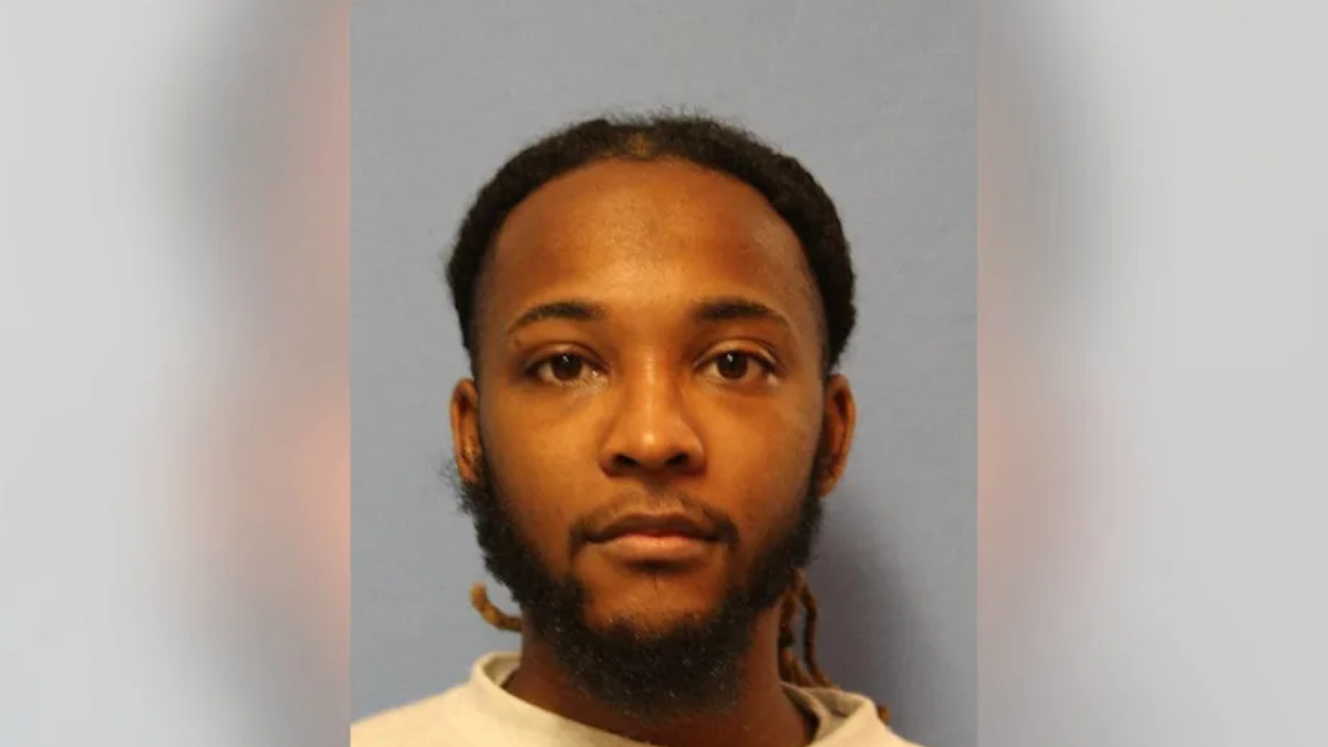 Mugshot-style portrait of a man with short dreadlocks and beard, neutral expression, gray-blue background.