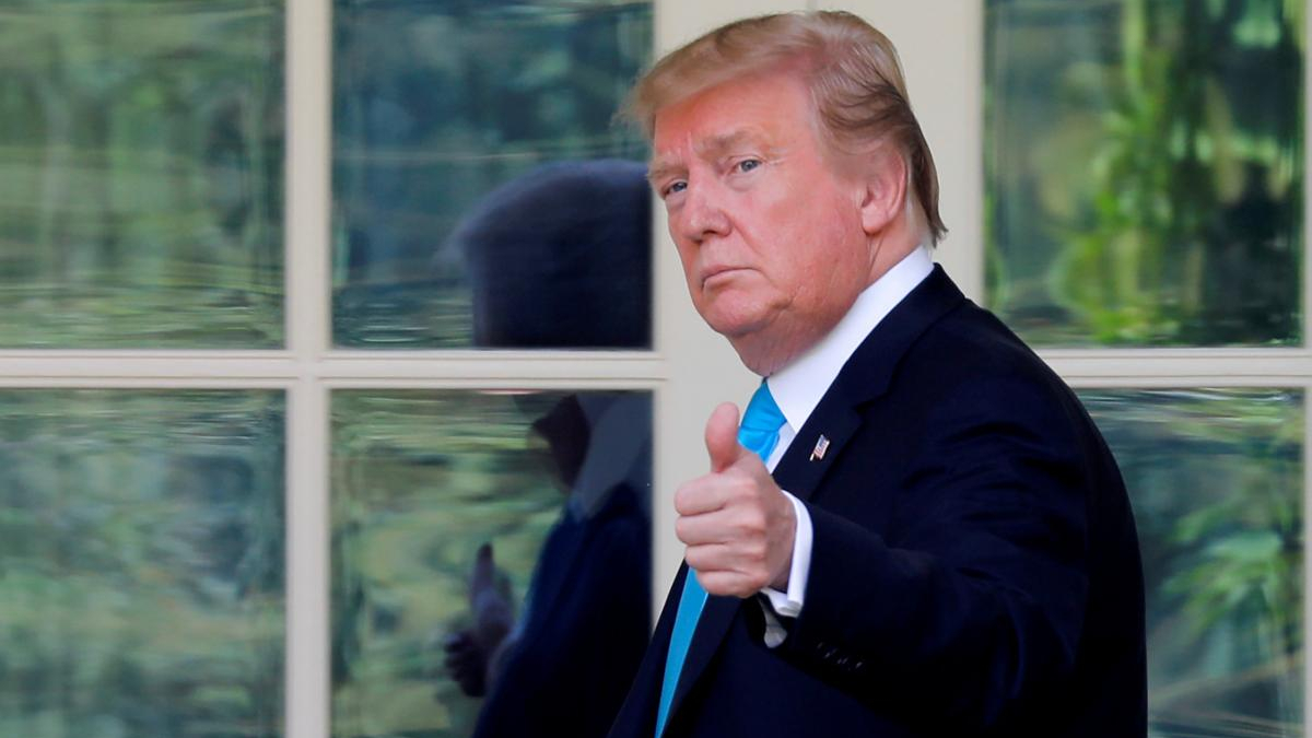 Man in a dark suit and bright blue tie giving a thumbs-up, facing the camera in front of glass doors/windows.