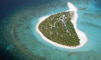Aerial view of a small tropical island with dense trees, white sand beaches, and a line of buildings along a central path. The surrounding turquoise water shows coral reefs nearby.