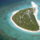 Aerial view of a small tropical island with dense trees, white sand beaches, and a line of buildings along a central path. The surrounding turquoise water shows coral reefs nearby.