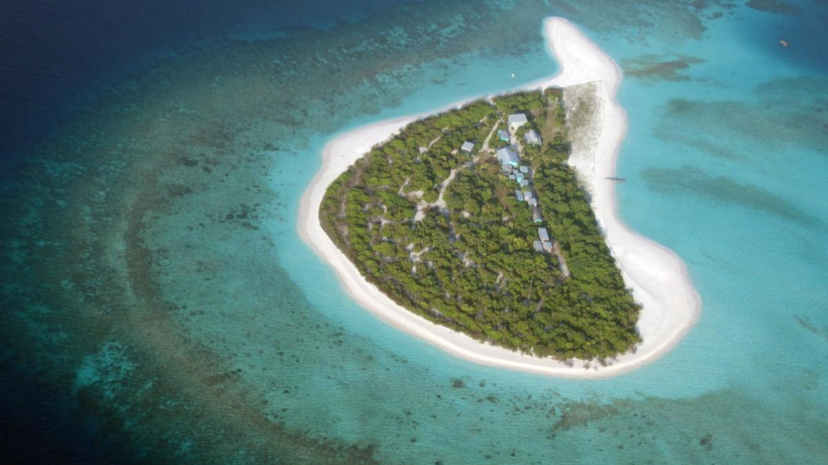 Aerial view of a small tropical island with dense trees, white sand beaches, and a line of buildings along a central path. The surrounding turquoise water shows coral reefs nearby.