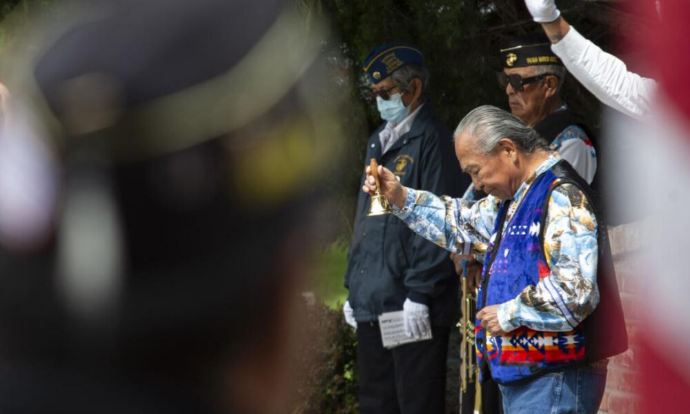 An elderly man in a colorful vest rings a small brass bell during a ceremonial gathering, with uniformed veterans nearby.