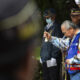 An elderly man in a colorful vest rings a small brass bell during a ceremonial gathering, with uniformed veterans nearby.