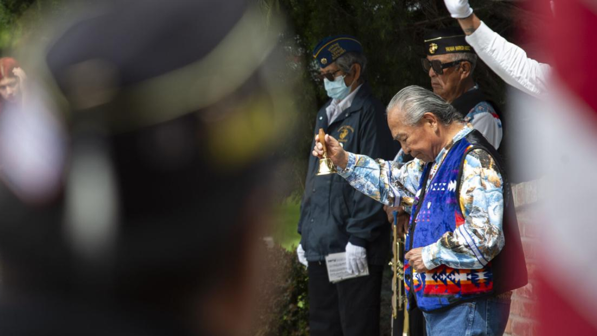 An elderly man in a colorful vest rings a small brass bell during a ceremonial gathering, with uniformed veterans nearby.
