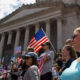 Protesters outside a large neoclassical government building, some holding signs and American flags.