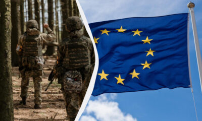Soldiers in camouflage gear walking through a forest, split by a diagonal white divider from a waving European Union flag against a blue sky; conveys military cooperation in Europe.